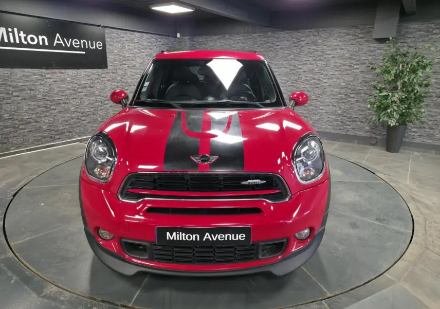 Vue avant d'une MINI Countryman John Cooper Works rouge avec bandes noires sport et calandre siglée, en intérieur showroom.