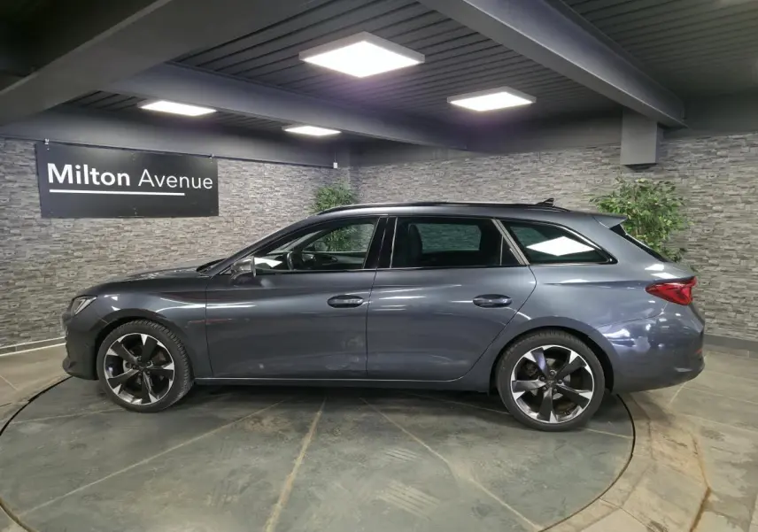 Vue de profil côté gauche d'une CUPRA Leon Sportstourer gris magnétique avec jantes alliage 18 pouces dans un showroom.