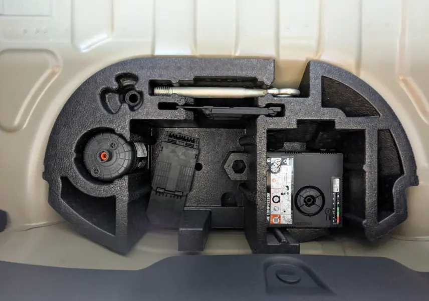 Vue rapprochée du compartiment de rangement sous le coffre de la Citroën E-C3 113 MAX gris Mercury, montrant l'outillage et compresseur.