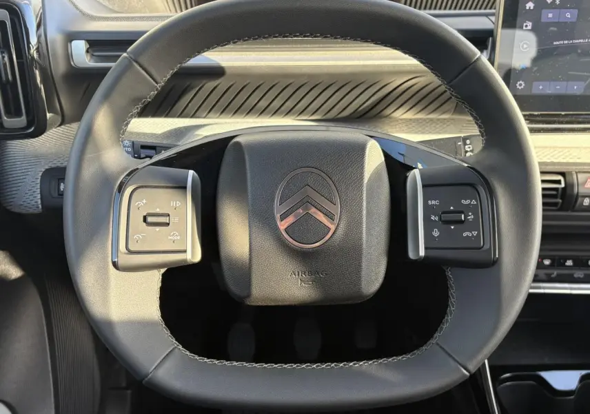 Vue rapprochée du volant noir de la Citroën C3 2025 avec commandes intégrées et tableau de bord moderne en arrière-plan.