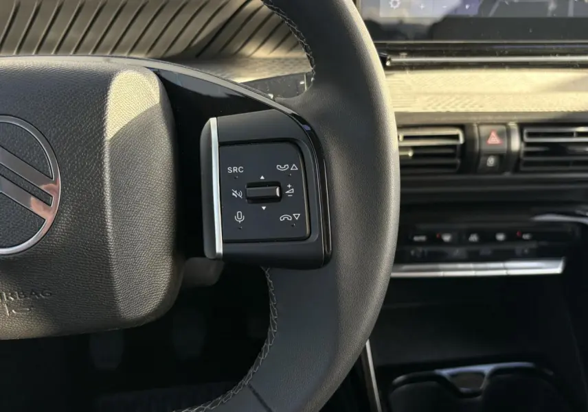 Gros plan sur la commande gauche du volant noir de la Citroën C3 2025 avec tableau de bord et écran tactile en arrière-plan.