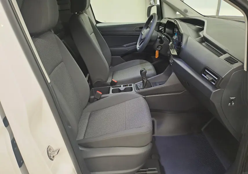 Vue intérieure côté conducteur du Volkswagen Caddy Cargo Maxi 2026 blanc, sièges gris et tableau de bord moderne avec boîte manuelle.