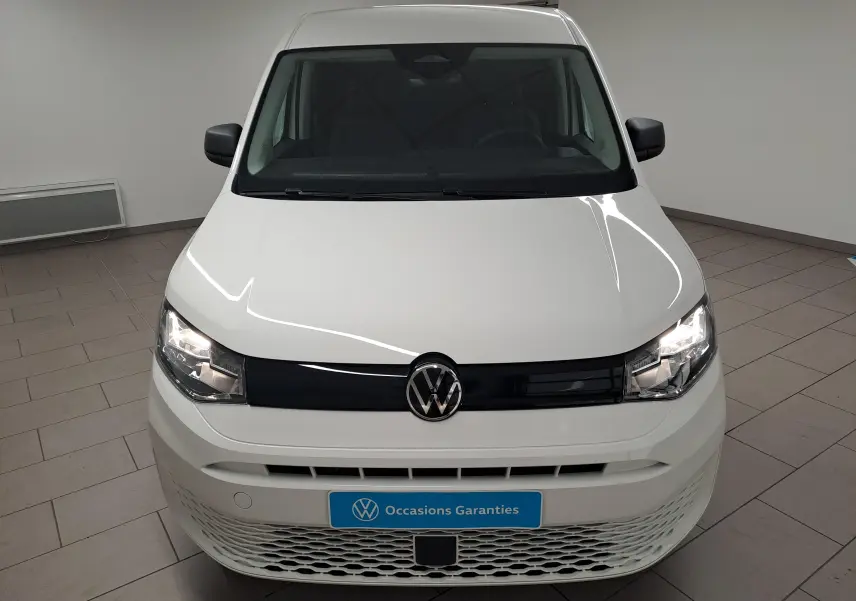 Vue frontale d’un Volkswagen Caddy Cargo Maxi blanc candy 2026 avec calandre noire et phares allumés en intérieur showroom.