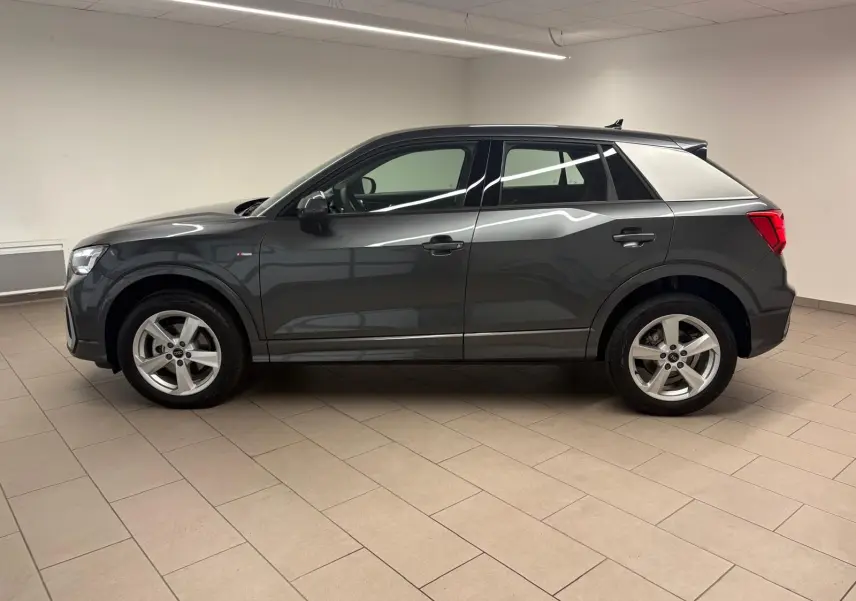 Audi Q2 35 TFSI S Line gris Daytona en profil gauche, avec jantes alliage et toit noir contrasté dans un intérieur neutre.