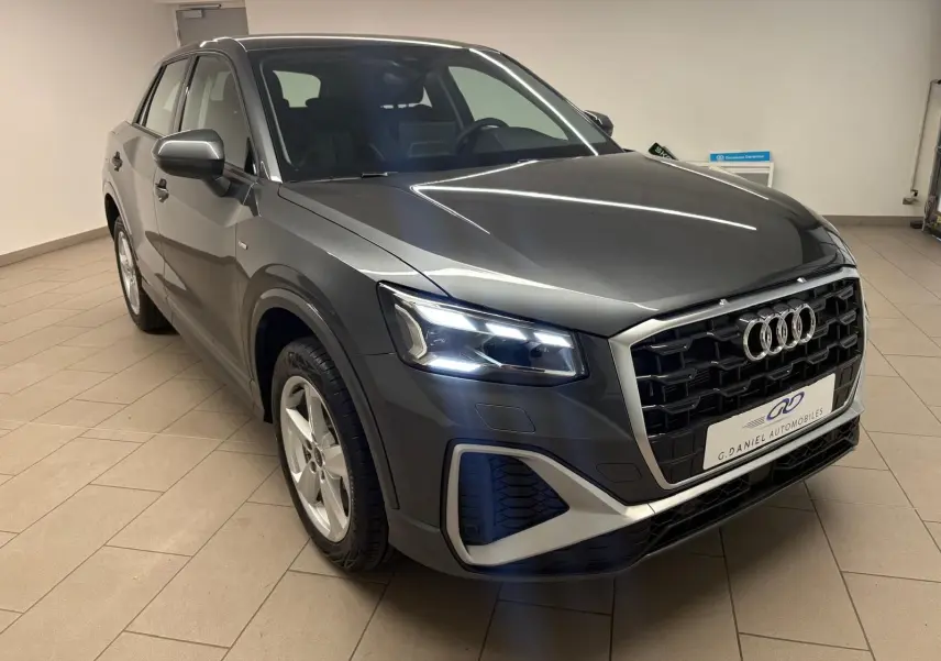 Audi Q2 35 TFSI S Line gris Daytona en 3/4 avant droit, avec calandre noire et phares LED allumés.