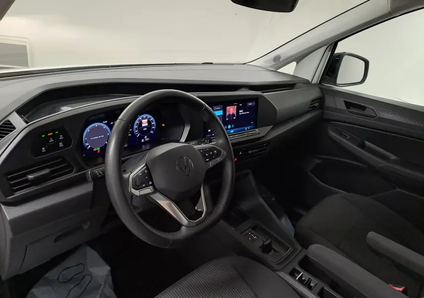 Vue intérieure côté conducteur du Volkswagen Caddy 2025, tableau de bord noir avec écran tactile et volant cuir multifonction.