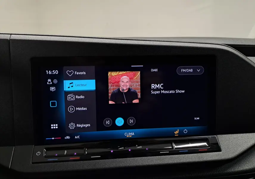 Écran tactile central du Volkswagen Caddy blanc 2025 affichant la radio RMC et les commandes climatisation.