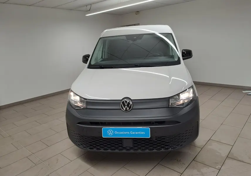 Vue avant d'un Volkswagen Caddy blanc Candy 2025 avec pare-chocs noir mat et phares allumés en intérieur.