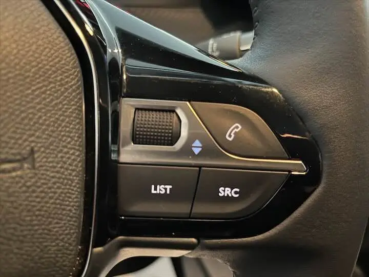 Gros plan sur les commandes gauche du volant noir du Peugeot 208 gris Artense, avec boutons LIST et SRC visibles.
