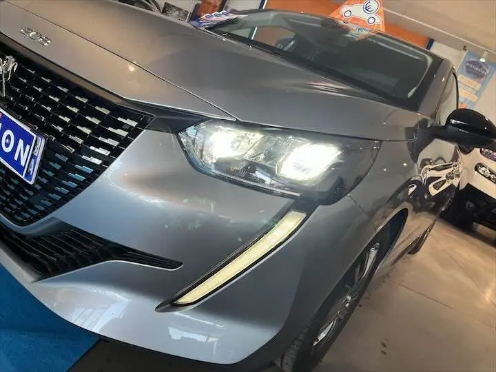 Vue 3/4 avant droit d'une Peugeot 208 gris Artense métal avec phares allumés en intérieur showroom.