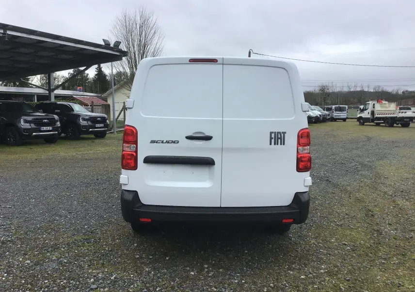 Vue arrière d’un FIAT Scudo Fourgon blanc 2024 avec portes battantes et feux arrière rouges sur un terrain extérieur.