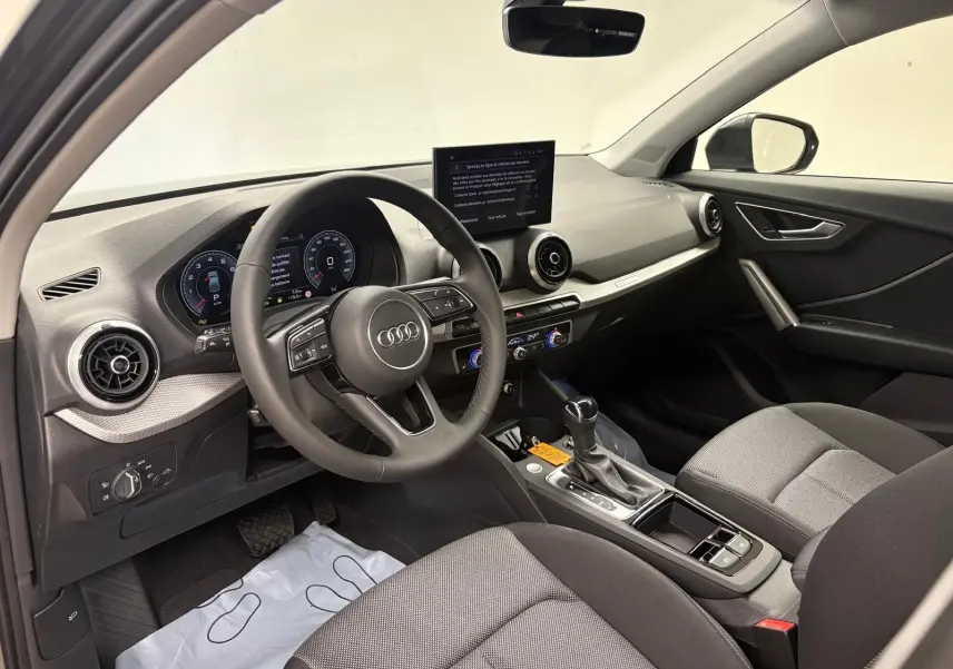 Vue intérieure côté conducteur de l'Audi Q2 35 TFSI S Line 2026, avec tableau de bord moderne et sièges tissu gris.