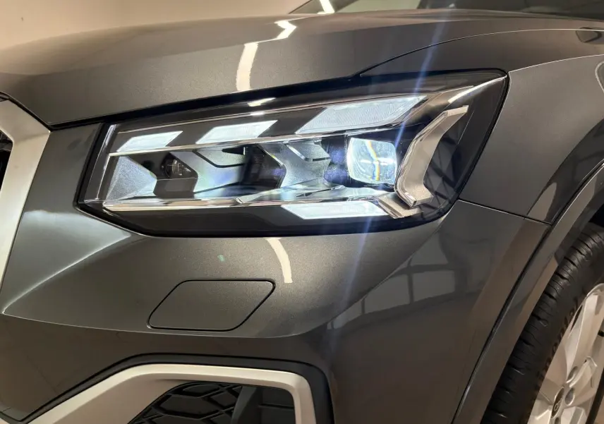 Gros plan sur le phare avant droit d’un Audi Q2 35 TFSI S Line gris Daytona métallisé, avec détails lumineux et carrosserie brillante.