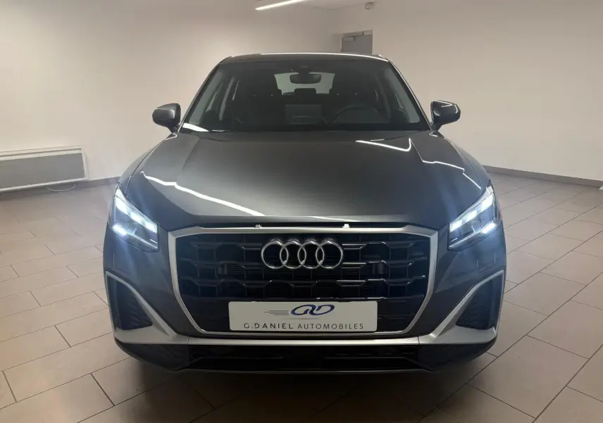 Vue frontale d’un Audi Q2 35 TFSI S Line gris Daytona avec phares allumés dans un showroom.