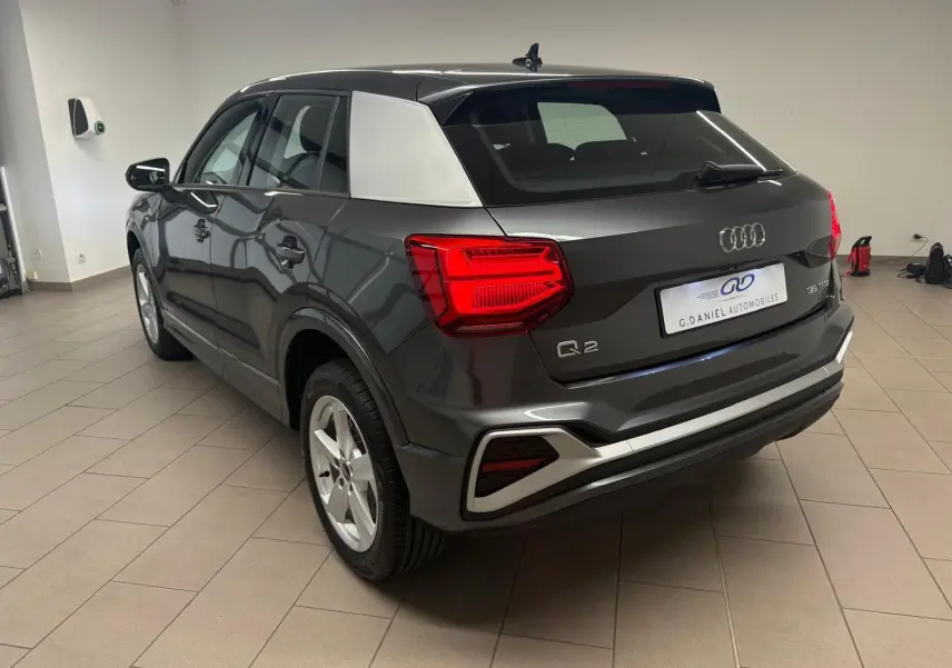 Audi Q2 35 TFSI S Line gris Daytona vu en 3/4 arrière droit, avec toit contrasté argent et feux arrière allumés.
