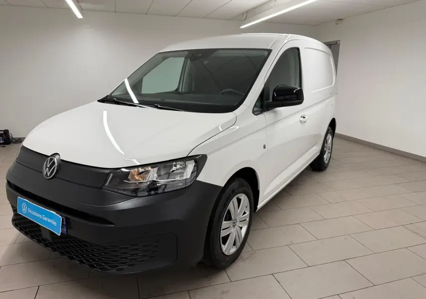 Vue 3/4 avant droit du Volkswagen Caddy blanc Candy avec pare-chocs noir mat et jantes acier en intérieur showroom.