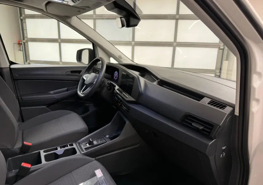 Vue intérieure côté conducteur du Volkswagen Caddy blanc Candy 2025, tableau de bord et console centrale noirs.