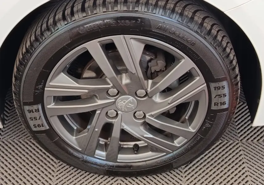 Gros plan sur la roue avant droite d'une Peugeot 208 blanche, avec jante alliage grise et pneu toutes saisons.