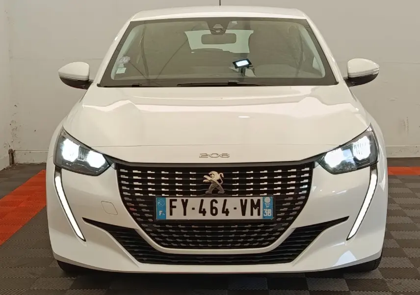 Vue avant d'une Peugeot 208 blanche de 2021 avec phares LED allumés et calandre noire distinctive.