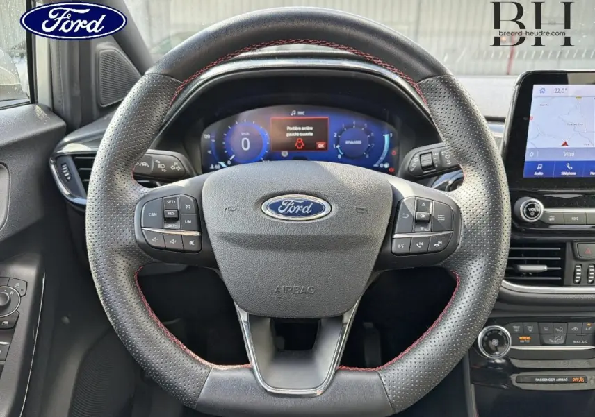 Vue intérieure centrée sur le volant perforé avec surpiqûres rouges du Ford Puma 2024 gris Solar Métallisée, tableau de bord digital visible.