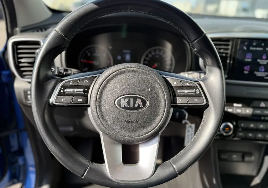 Vue rapprochée du volant noir du KIA Sportage bleu 2021 avec commandes intégrées et tableau de bord en arrière-plan.