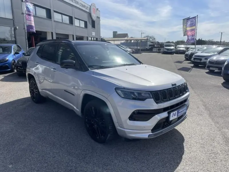 Jeep Compass gris métal vu en 3/4 avant droit, avec calandre noire distinctive et jantes noires, stationné en extérieur.