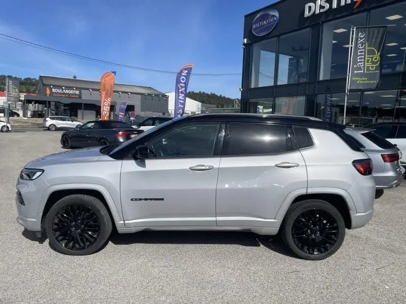 Vue de profil côté gauche d'un Jeep Compass gris métal 2023 avec jantes noires et toit noir, stationné devant un bâtiment commercial.