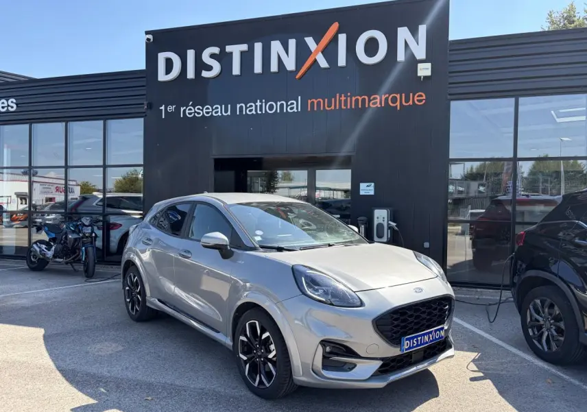 Ford Puma gris métal en 3/4 avant droit, avec jantes alliage noires et calandre ST-Line visible devant un showroom Distinxion.