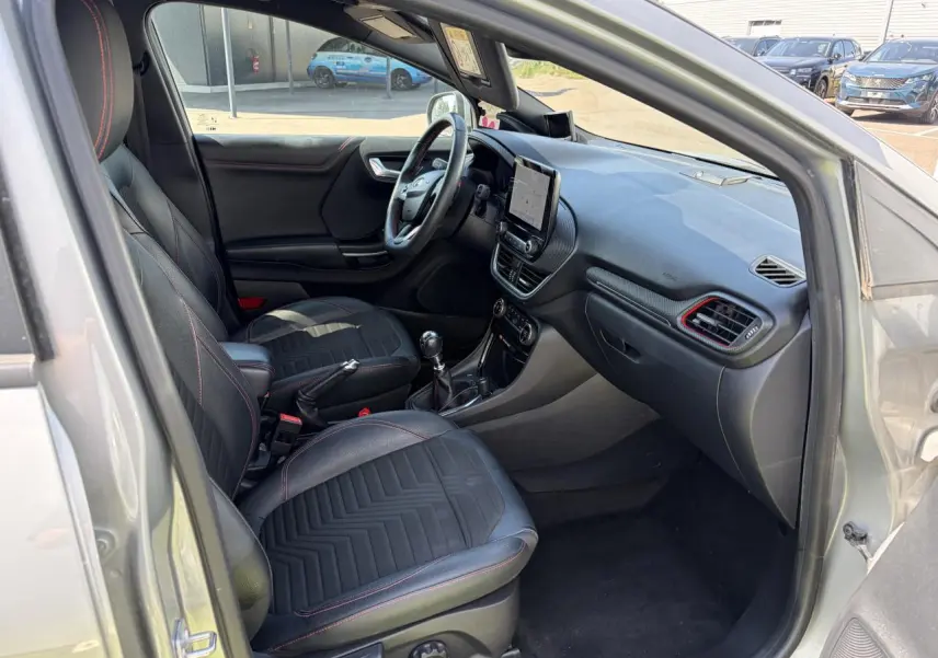 Intérieur avant du Ford Puma gris métal, vue côté conducteur, sièges noirs avec surpiqûres rouges et tableau de bord moderne.