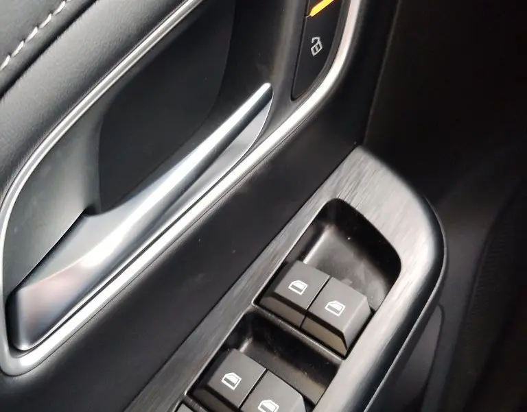 Gros plan sur la commande des vitres et la poignée intérieure côté gauche du MG ZS 1.5 l Hybrid+ 197 Luxury en finition noire.