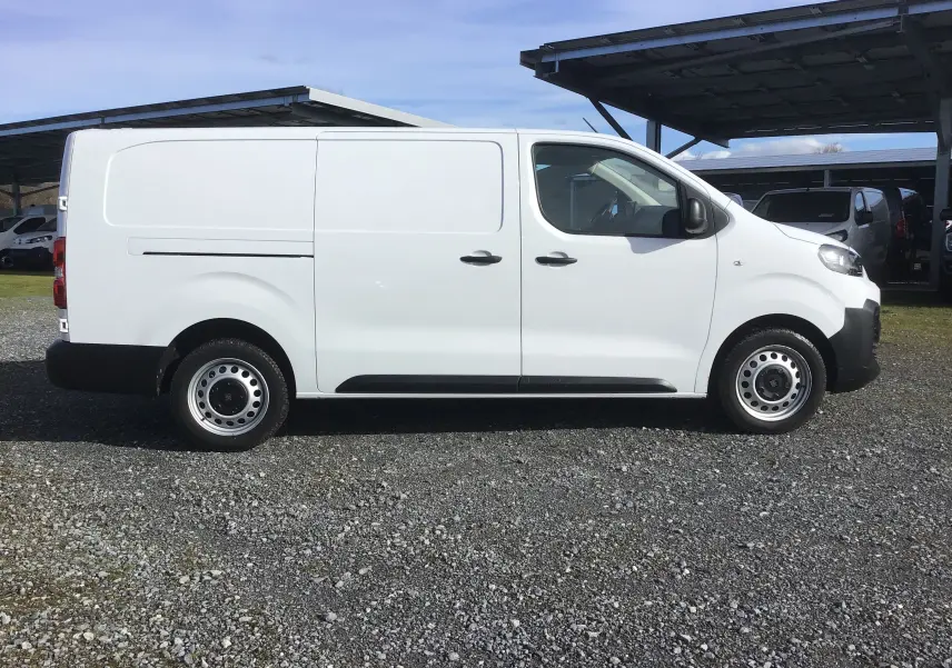 Vue de profil côté gauche d'un FIAT Scudo Fourgon blanc 2024 avec garde au sol réhaussée sur un sol gravillonné.