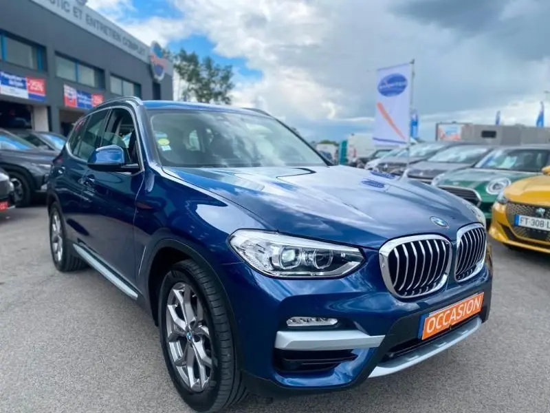 BMW X3 X-DRIVE 20 DA bleu métal en 3/4 avant droit, avec calandre chromée et plaque "OCCASION" visible.