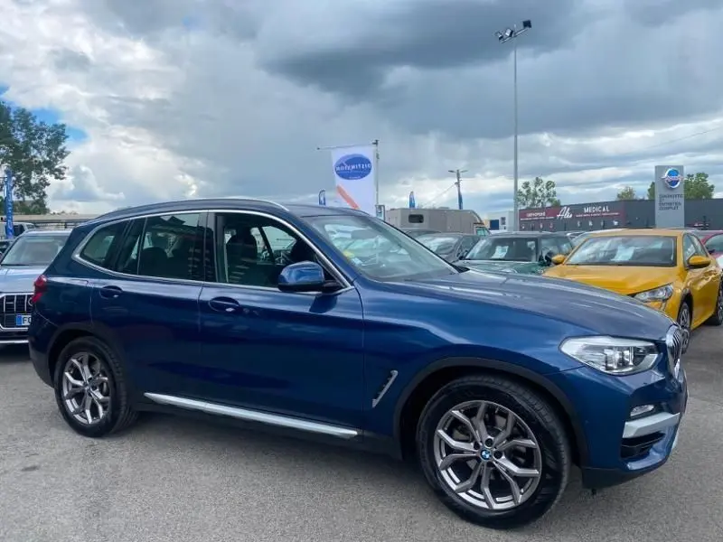 BMW X3 X-DRIVE 20 DA bleu métal en vue 3/4 avant droit, avec jantes alliage et détails X-Line visibles.