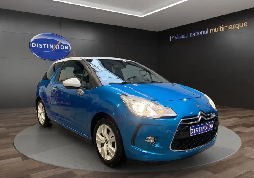 Citroën DS3 bleu avec toit blanc en 3/4 avant droit, phare allumé et jantes blanches visibles en intérieur showroom.