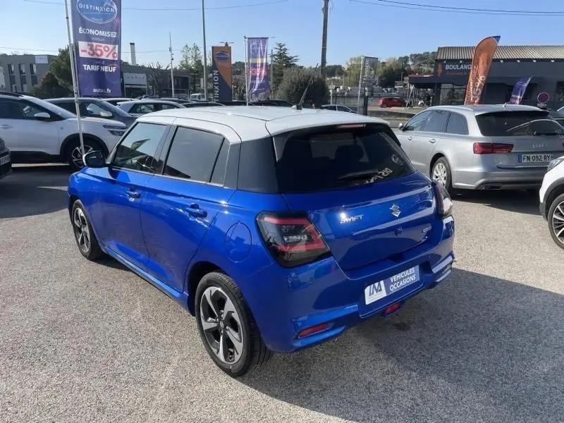 Vue 3/4 arrière droite d'une Suzuki Swift 2024 bleu métal avec toit blanc et feux arrière fumés sur parking extérieur.
