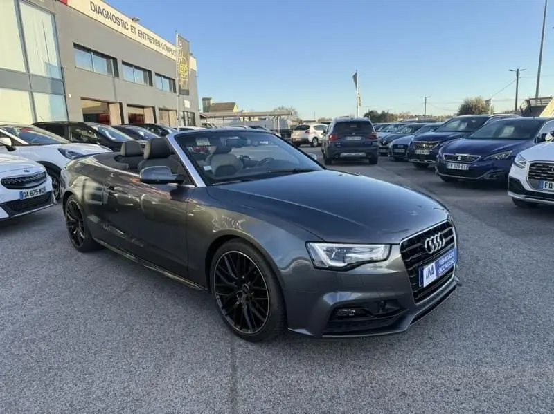 Audi A5 Cabriolet gris métal vue 3/4 avant avec capote ouverte et jantes noires dans un parking extérieur.