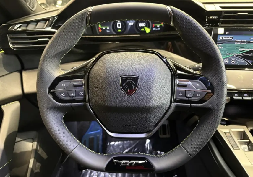 Vue rapprochée du volant GT en cuir noir avec surpiqûres vert Adamite du Peugeot 308 2026, tableau de bord digital visible.