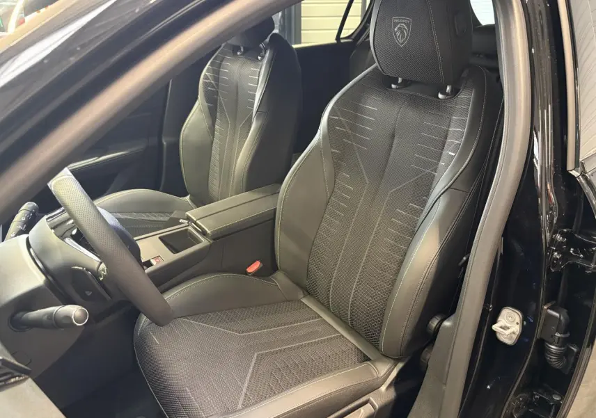 Vue intérieure côté conducteur de la Peugeot 308 GT 2026 noire métallisée, sièges sport avec logo Peugeot brodé.