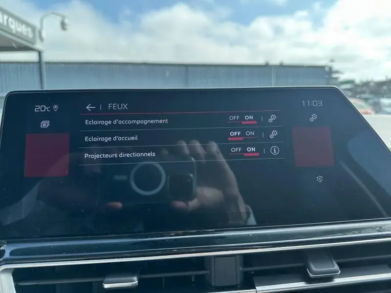 Écran tactile du tableau de bord du Citroën C5 Aircross 2025 affichant les réglages des feux, avec reflet de la main et de l'appareil photo.