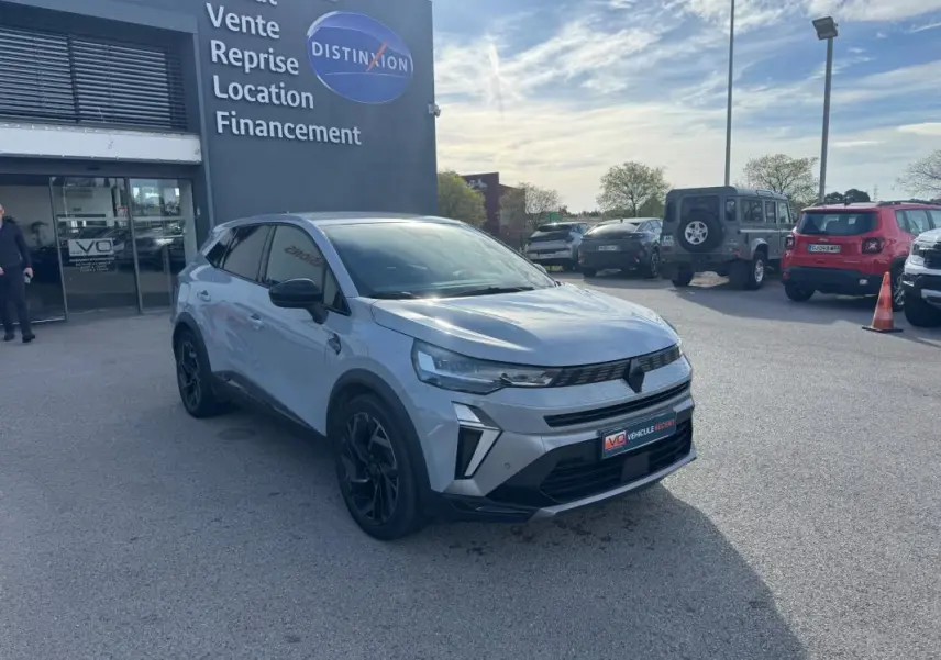 Renault Symbioz gris clair en 3/4 avant droit, jantes alliage noires de 19 pouces, devant un concessionnaire.