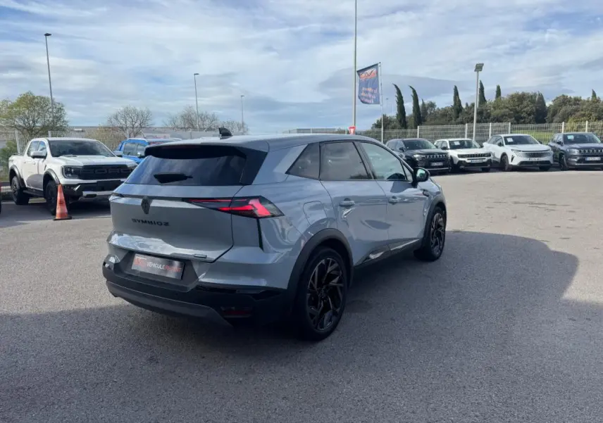 Vue 3/4 arrière droite d'un Renault SYMBIOZ gris clair avec jantes alliage noires et antenne requin sur parking.