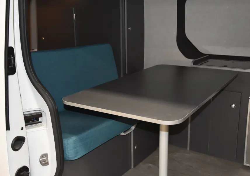 Coin repas intérieur avec banquette bleue et table grise dans un Renault Trafic Combi blanc, porte latérale ouverte.
