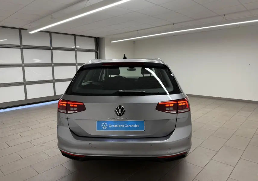 Vue arrière d'une Volkswagen Passat SW 2.0 TDI 122 DSG7 Business gris pyrite avec feux allumés en intérieur showroom.