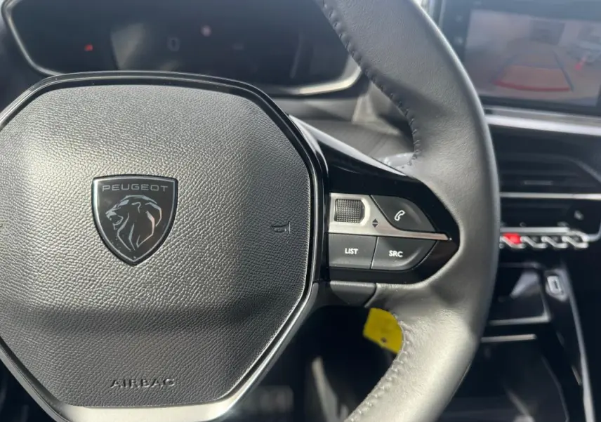 Vue rapprochée du volant noir de la Peugeot 2008 2024 avec commandes intégrées et tableau de bord numérique en arrière-plan.