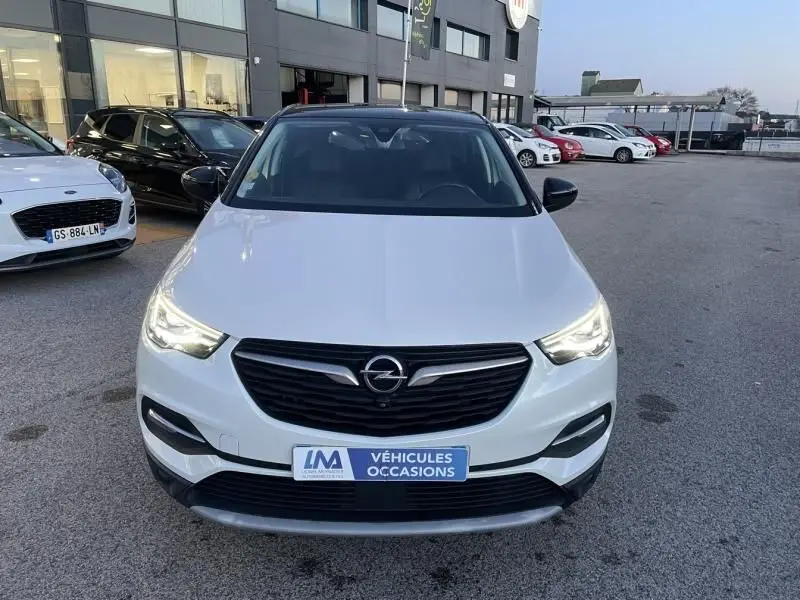 Vue avant d’un Opel Grandland X blanc avec toit noir, phares allumés, sur parking extérieur devant un bâtiment.