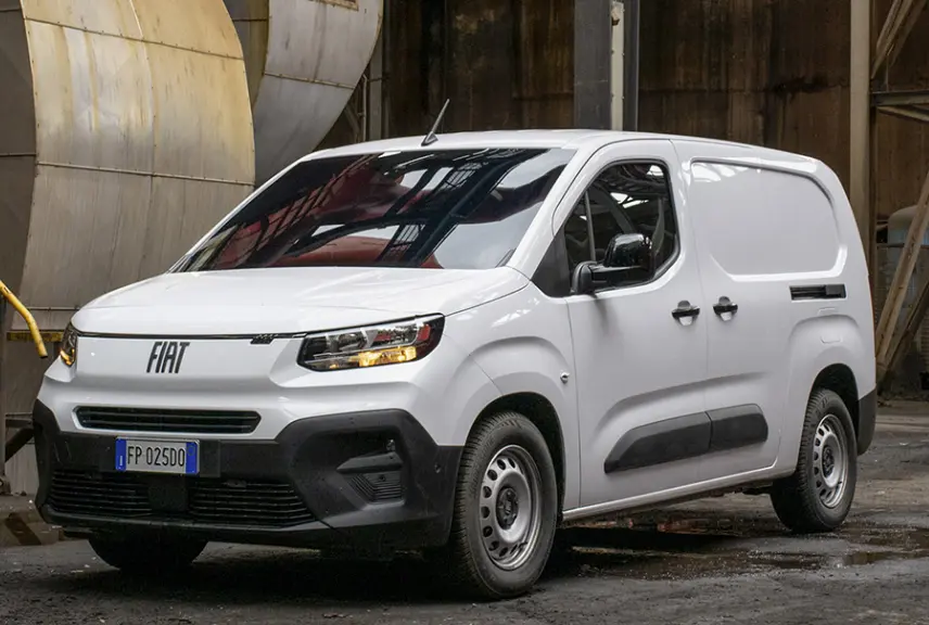 Fiat Doblo fourgon blanc vu en 3/4 avant droit dans un environnement industriel, avec calandre et phares allumés.