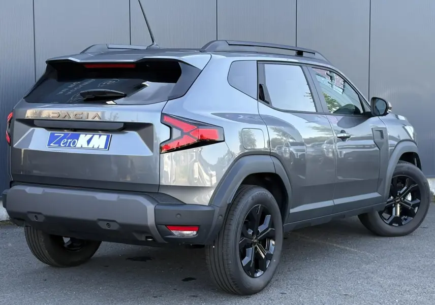 Vue 3/4 arrière droit du Dacia Duster 2026 gris schiste avec jantes alliage noires et barres de toit longitudinales.