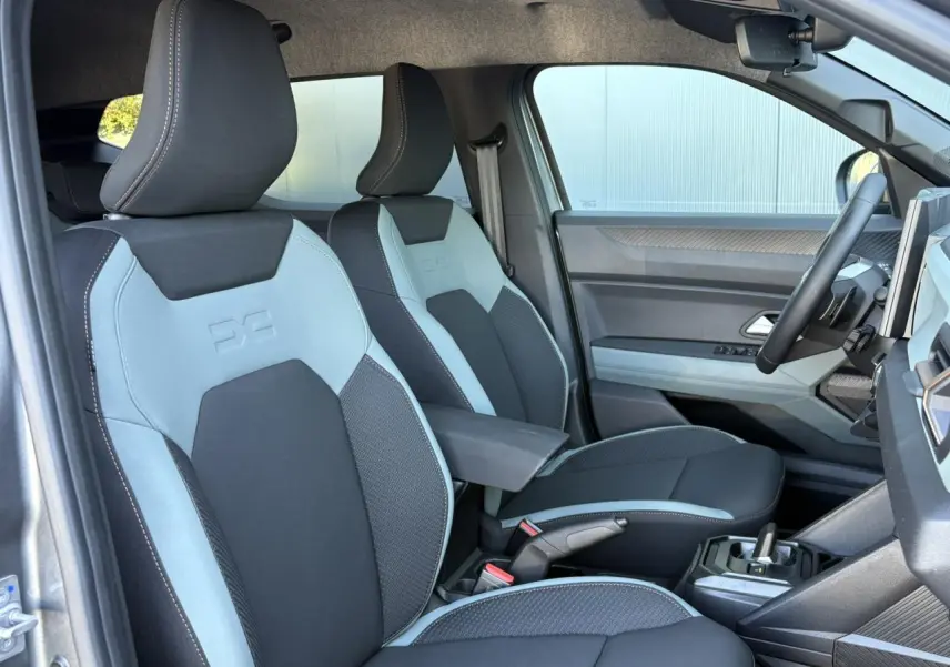 Vue intérieure côté conducteur du Dacia Duster 2026 gris Schiste, sièges bi-ton gris clair et noir avec accoudoir central.