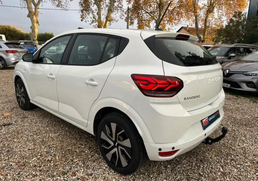 Vue 3/4 arrière droite d'une Dacia Sandero blanche 2024 avec feux arrière LED et attelage de remorque visible.