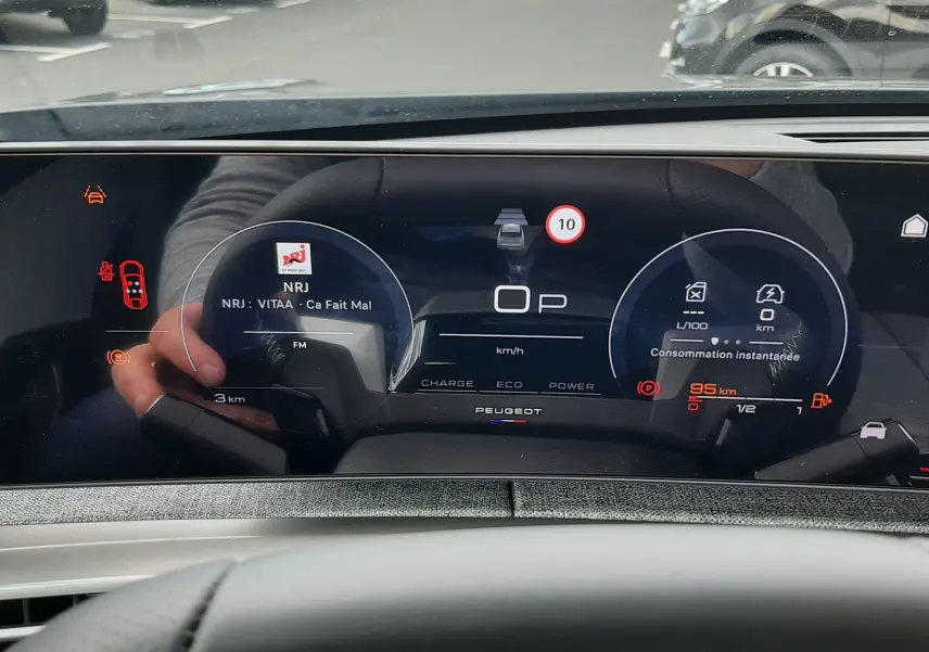 Vue intérieure du tableau de bord numérique du Peugeot 3008 hybride, affichant la vitesse, la radio NRJ et les indicateurs de consommation.